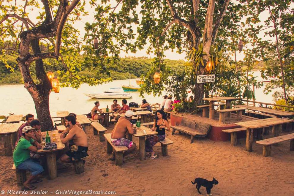 Imagem turistas em mesas e cadeiras de madeiras no meio das árvores aguardando entardecer-Caraiva.